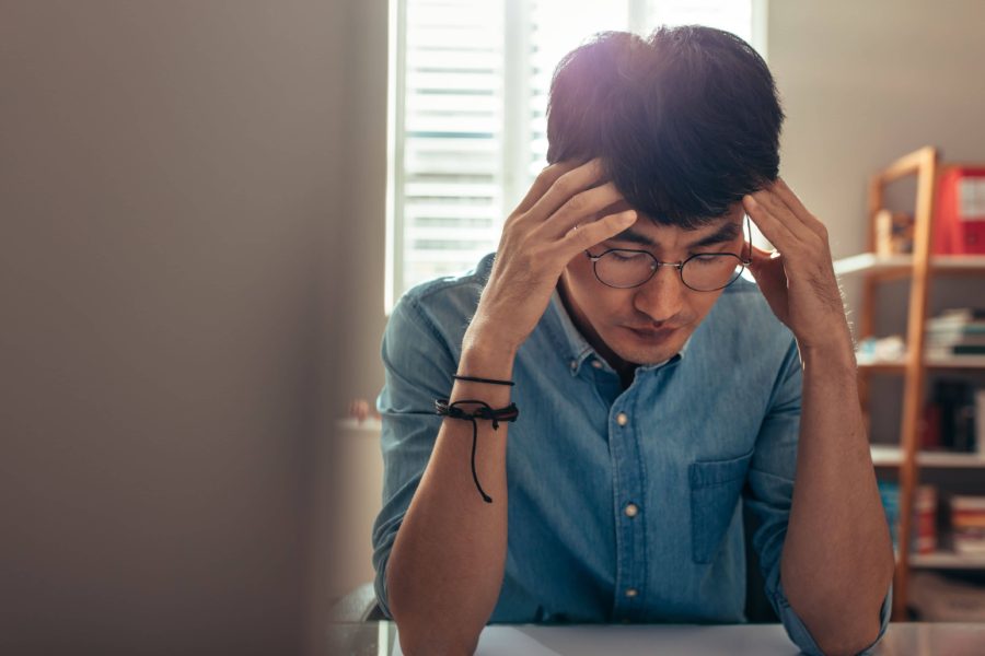 A man with glasses and a blue shirt touches his temples and looks down with concern.