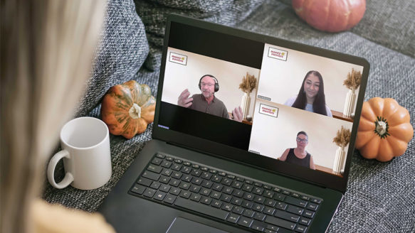 A woman watches a Money Mentors Financial Literacy Month webinar on her laptop, featuring three staff members on screen sharing budgeting and money management advice. Pumpkins and a coffee mug sit nearby, reflecting the November season.