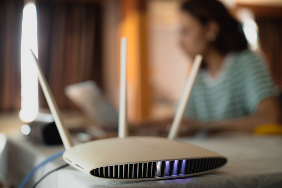 A router with antennas in the foreground and a person using a laptop in the background; save money on internet bills with tips from Money Mentors.