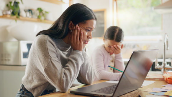 A stressed woman sitting at her kitchen table with her child, looking worried while using a laptop — a visual representation of financial burnout and stress.