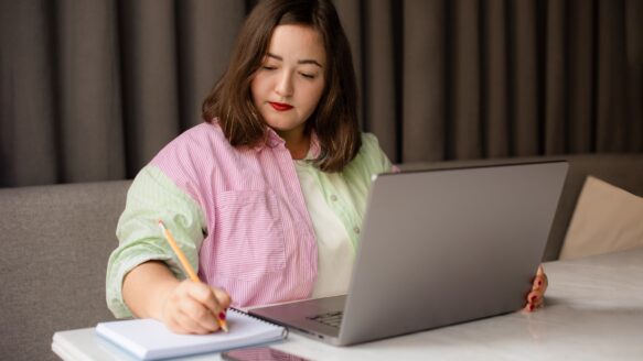 Focused female freelancer writing notes in notepad while working on laptop in cafe