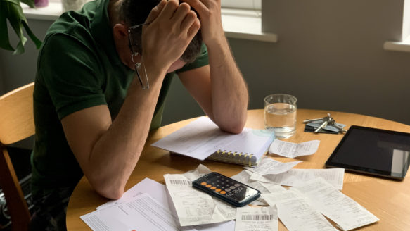 A man sits at a kitchen table with his head in his hands, surrounded by bills, receipts, and a calculator, stressed about rising household costs.