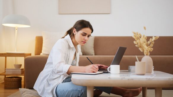 A young woman sits with a laptop and notebook, taking notes.