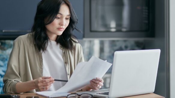 A woman reviews financial documents on a laptop at her kitchen table.