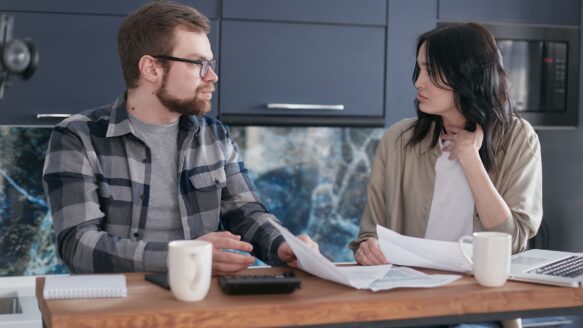 A couple sits at a kitchen table having a conversation over paperwork and coffee.