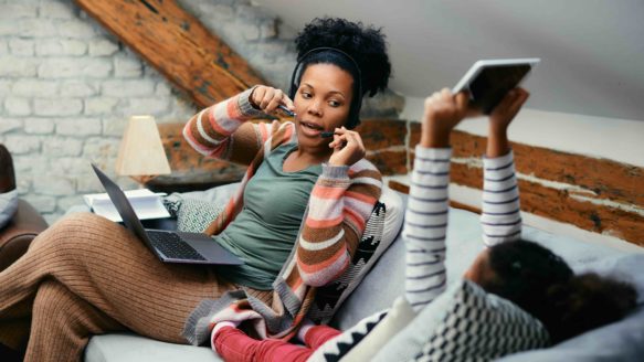 A multitasking mother in Alberta works from home during a side hustle shift while caring for her child, reflecting the pressure of rising living costs and burnout from juggling multiple jobs.