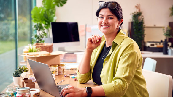 Smiling small business owner working on taxes at a home office, surrounded by shipping boxes and product jars.